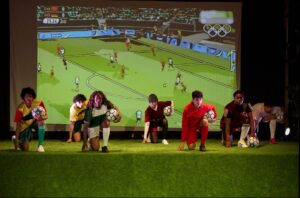 Jugendliche in Trikots knien auf einem grünen Rasen. Sie haben alle einen Fußball in der Hand. Auf einer großen Leinwand im Hintergrund ist ein Fußballspiel zu sehen.